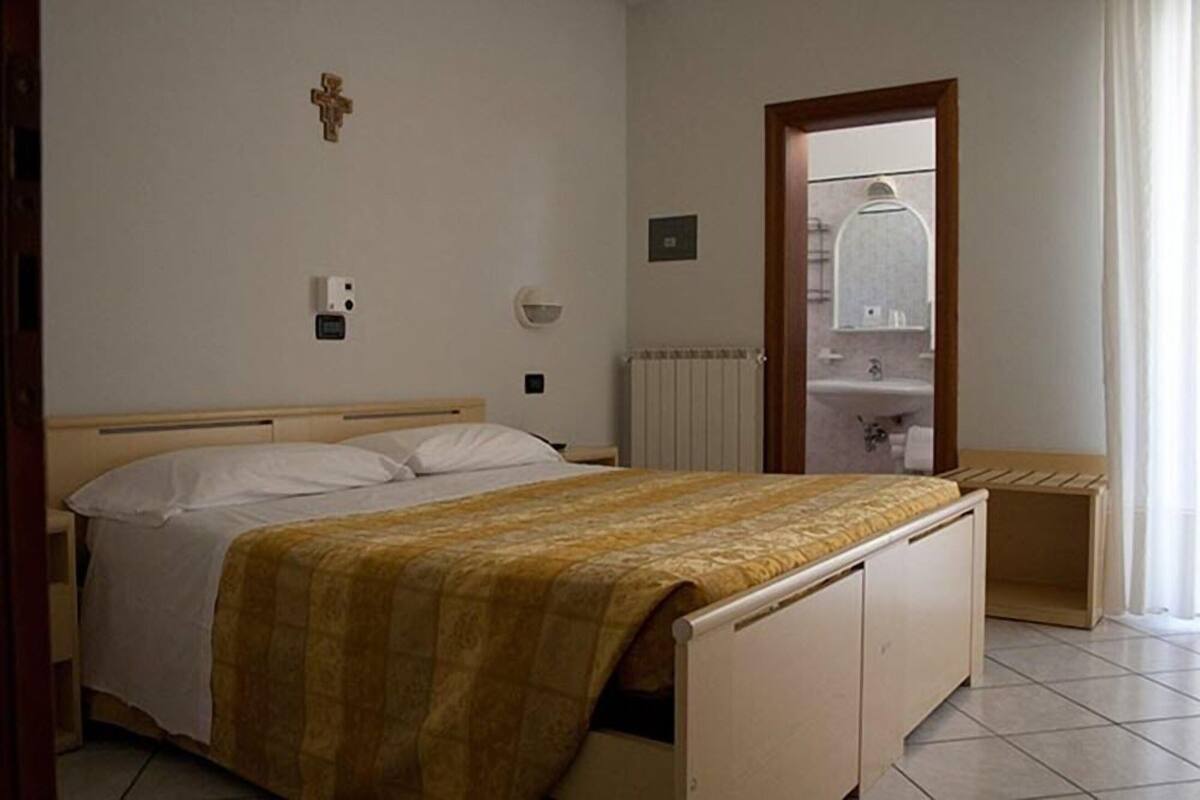 Un dormitorio con una cama, una mesita de noche, un radiador, un espejo y una puerta que da al baño.