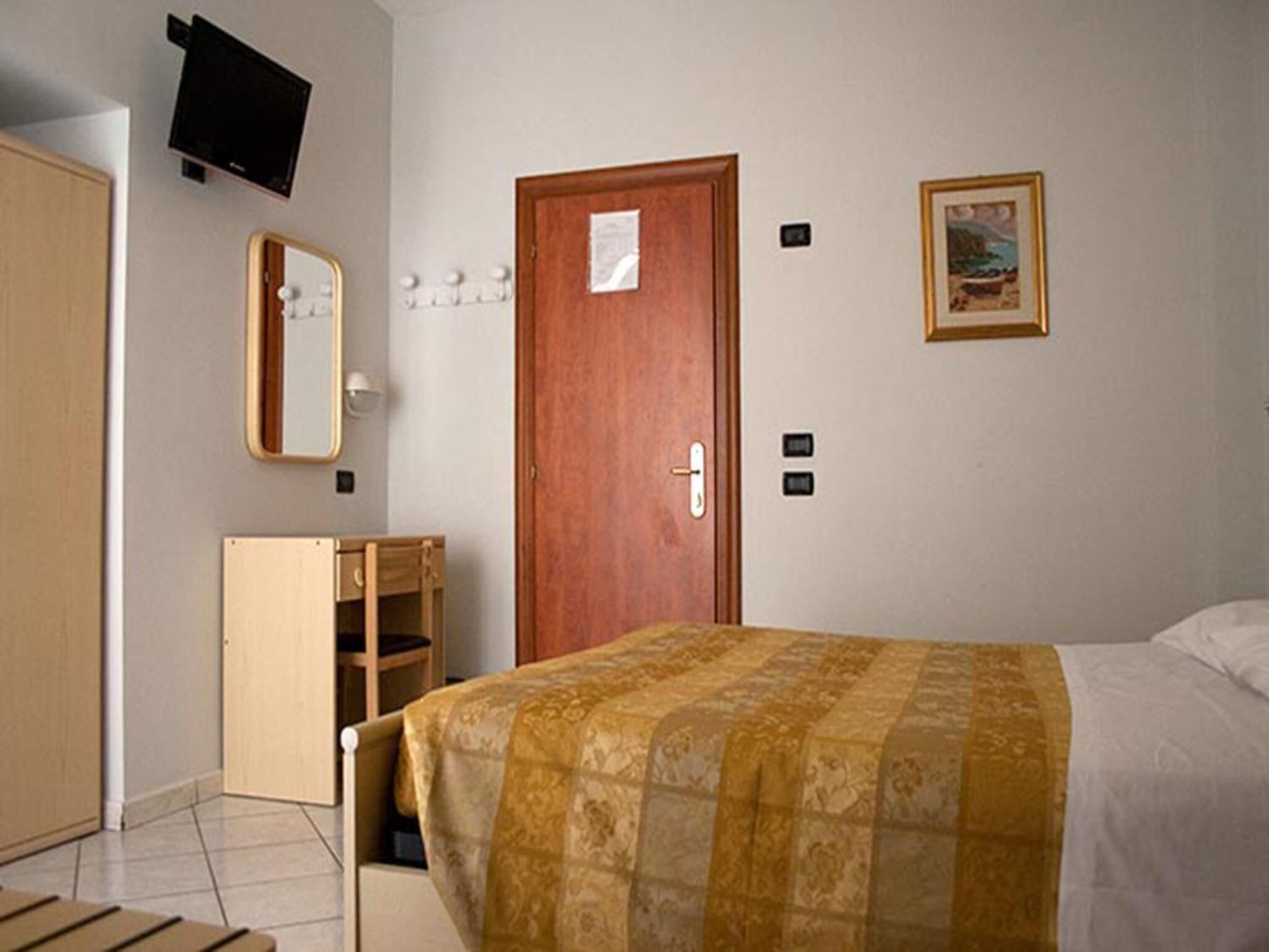 Un dormitorio con cama, un armario de madera, un espejo, una puerta y un televisor montado en la pared.