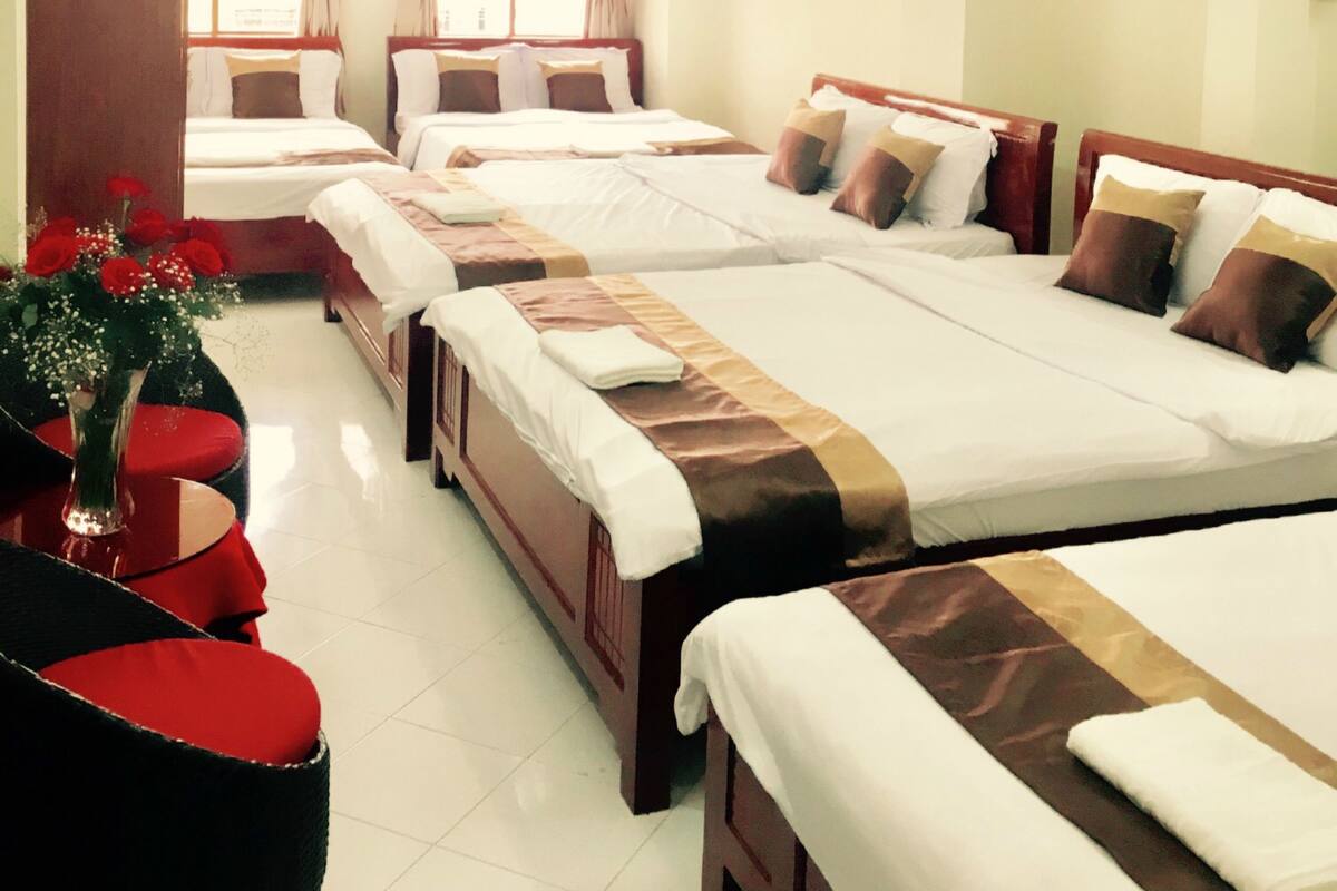 A hotel room with three beds, a red chair, a vase with flowers, and a painting on the wall.