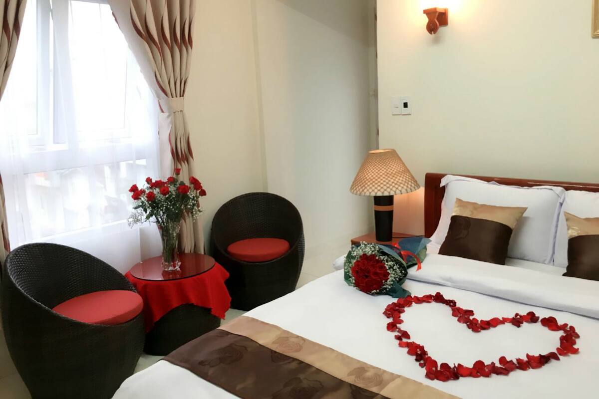 A hotel room with a bed, two armchairs, a small table with a vase of flowers, a lamp, and a bouquet of red roses.
