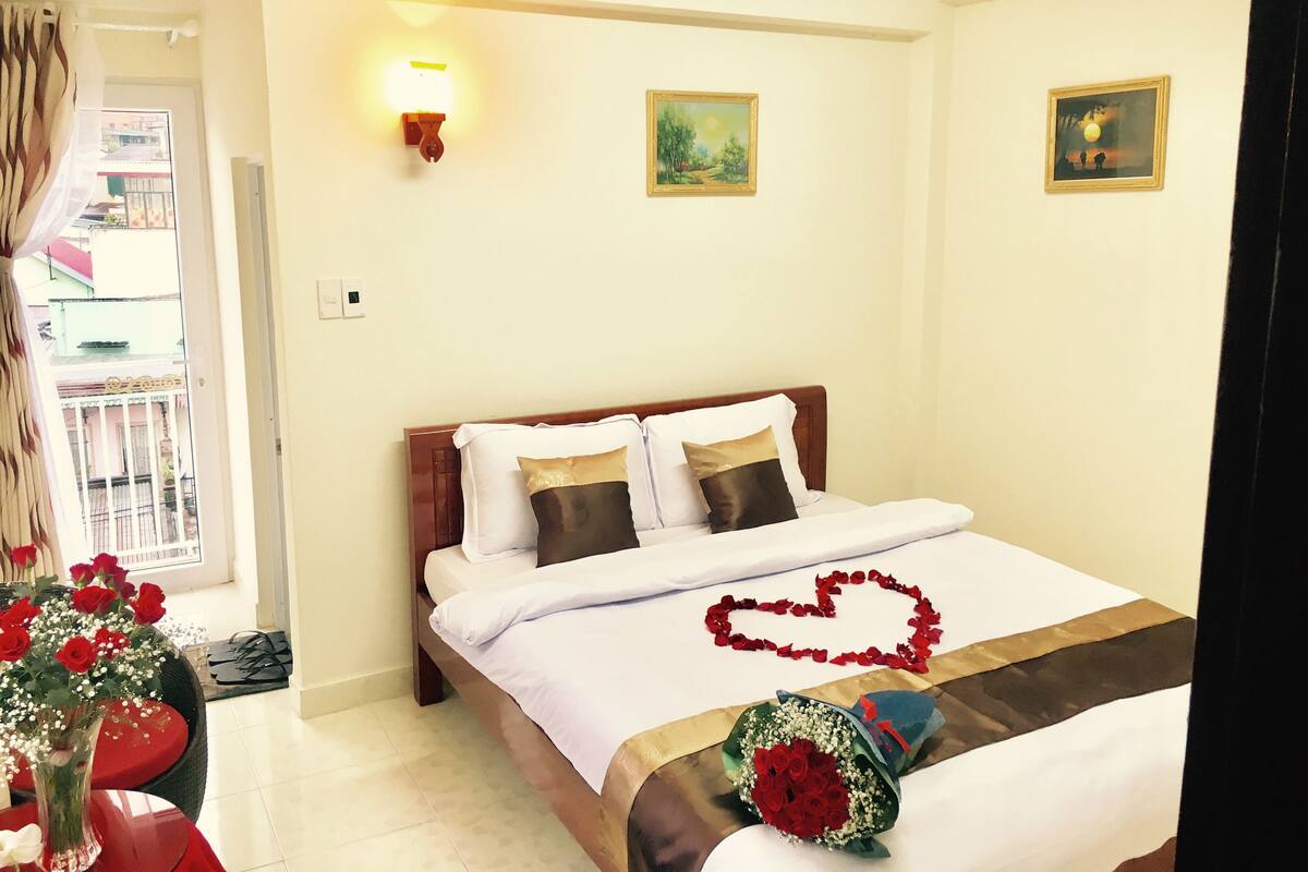 A hotel room with a bed, a nightstand, a vase with flowers, and a view of the outside balcony.