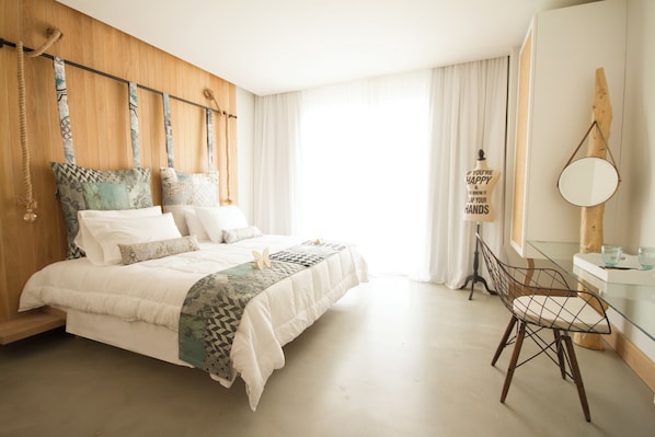 Une chambre à coucher avec une tête de lit en bois, un lit recouvert de linge de lit blanc, une table en verre avec une chaise, et une grande fenêtre avec des rideaux.