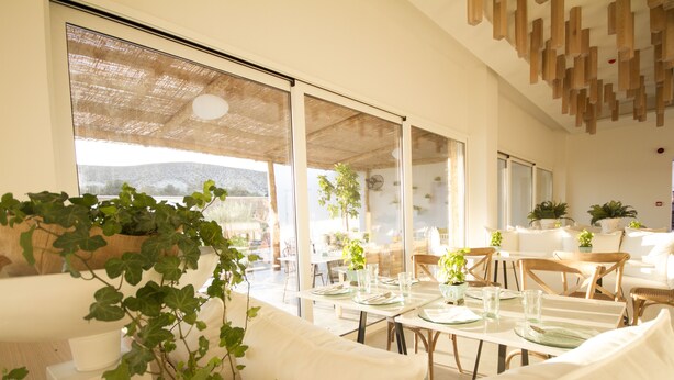 A bright, open-plan living area with a large window, a dining table set for two, and a sofa. - Aelia Wellness Retreat (Analiontas)