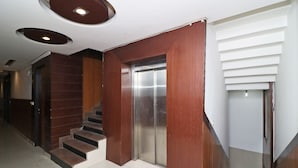 Um elevador moderno com acabamento metálico, uma escadaria de madeira com degraus escuros e uma parede branca com linhas horizontais.