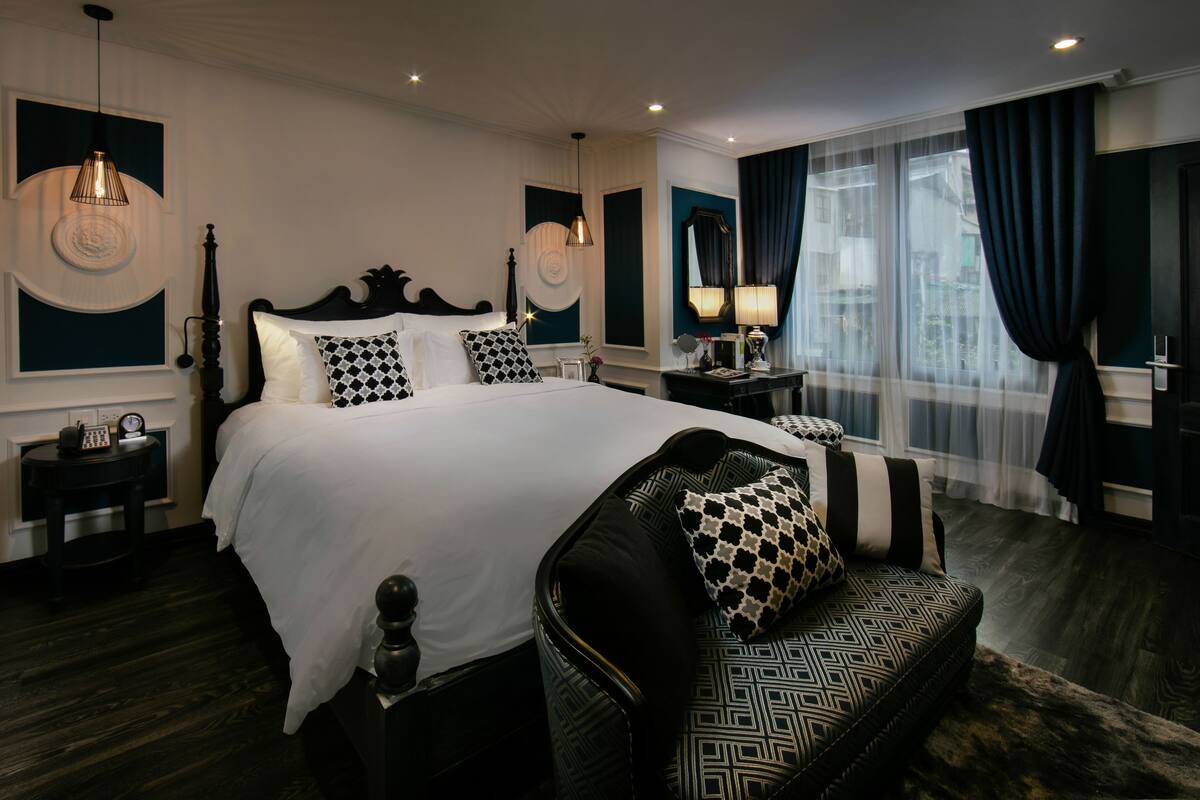 A modern hotel room with a large bed, a sofa, a nightstand, a lamp, and a view of the city through the window.