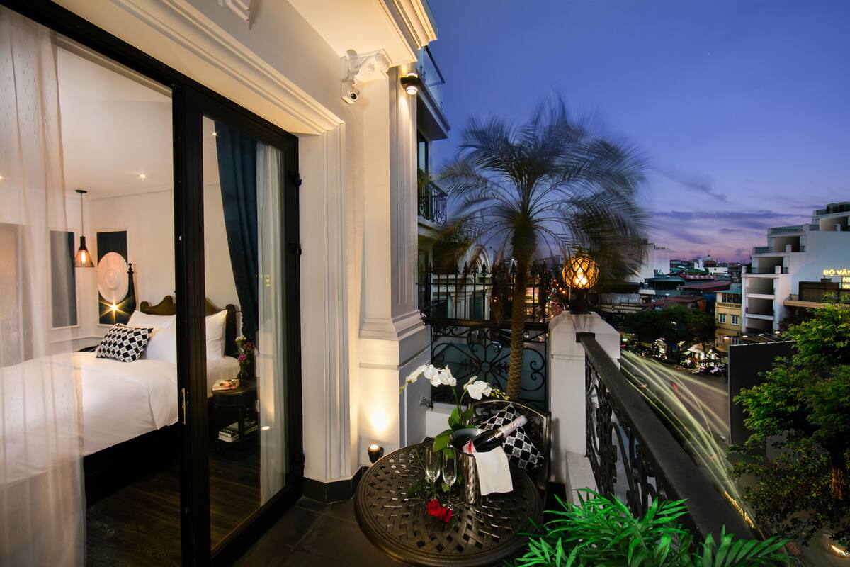 A hotel room with a bed, a small table with a bottle and flowers, a balcony with a palm tree, and a city view at dusk.