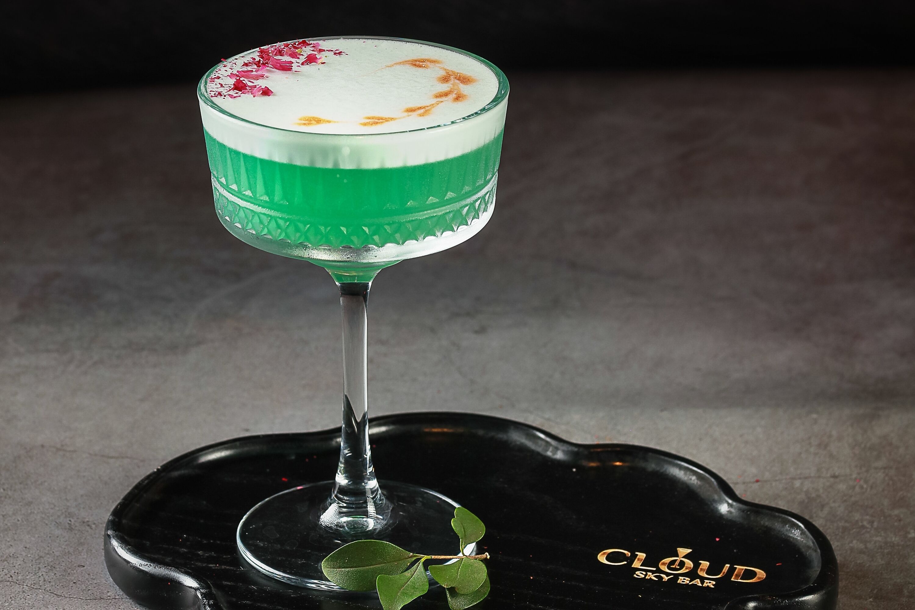 A cocktail glass with a green liquid and a white foam top, placed on a black tray with a leaf and the text "CLOUD SKY BAR".