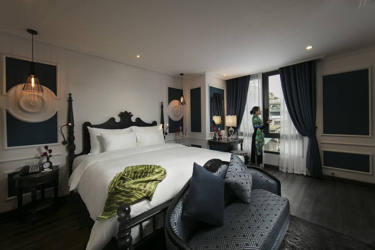 A hotel room with a large bed, a sofa, a nightstand with a lamp, and a view of the city through the window.