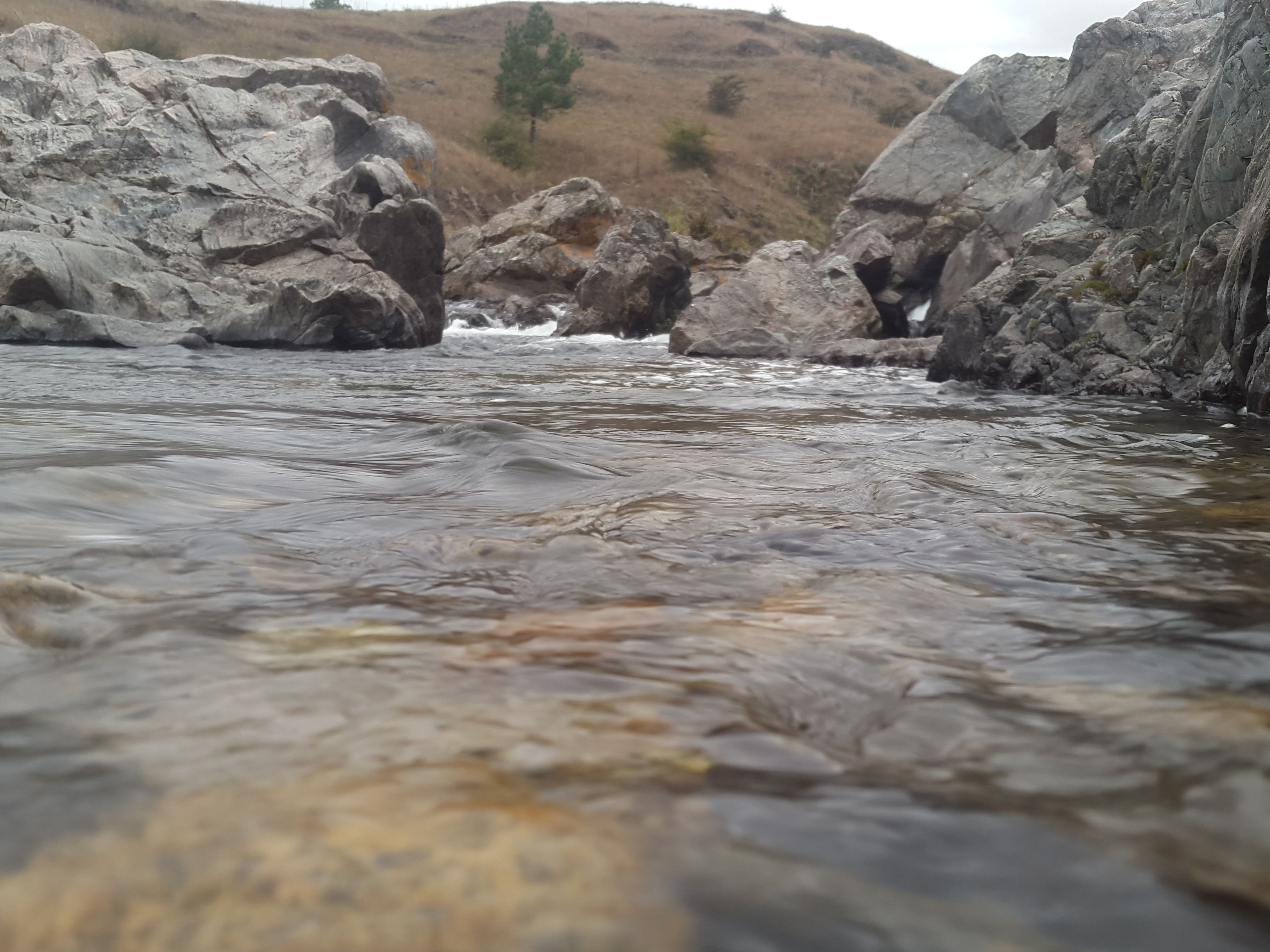 A river flowing between large rocks with a hill in the background.