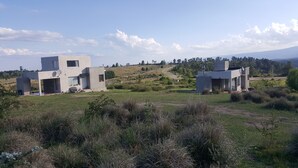 Two modern, minimalist houses with large windows and flat roofs, surrounded by a grassy field and a hilly landscape in the background. - Bernalina (La Cumbrecita)