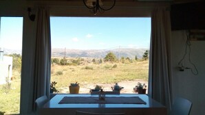 A room with a view of a grassy landscape and mountains. There is a table with a chair and a window with curtains. - Bernalina (La Cumbrecita)