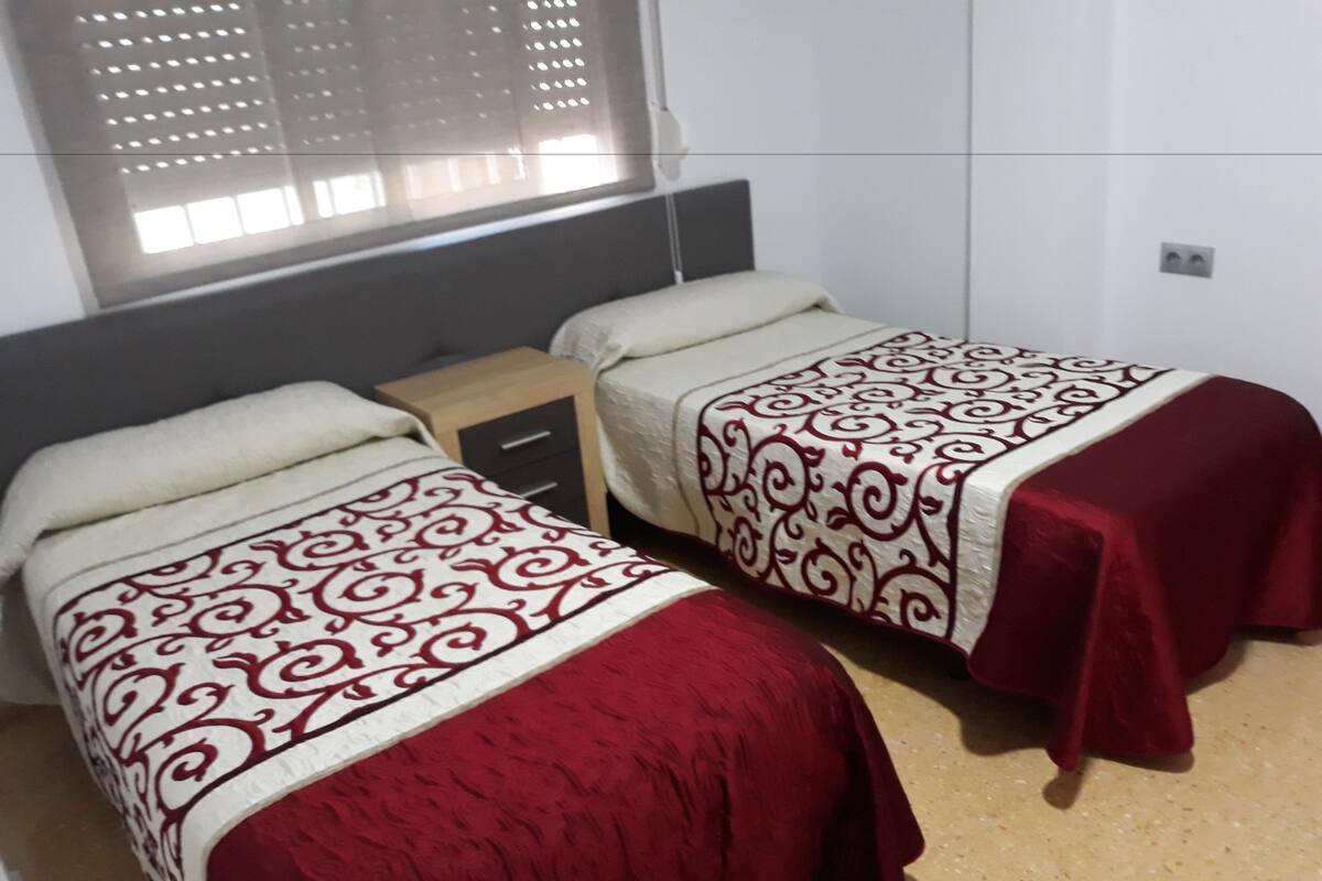 Dos camas individuales con colchas estampadas, una mesita de noche de madera y una ventana con persianas.