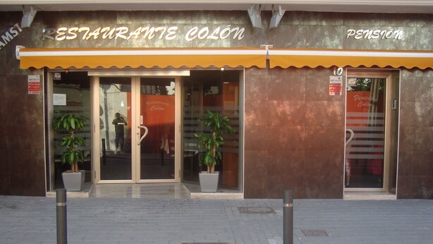 A restaurant with a yellow awning and glass doors. There are potted plants outside. - Pensión Colón (San Juan de Alicante)