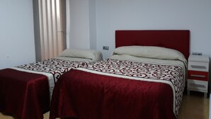 A room with two beds, each with a red headboard and patterned bedding. There is a window with blinds, an air conditioning unit on the wall, and a bedside table with drawers. - Pensión Colón (San Juan de Alicante)