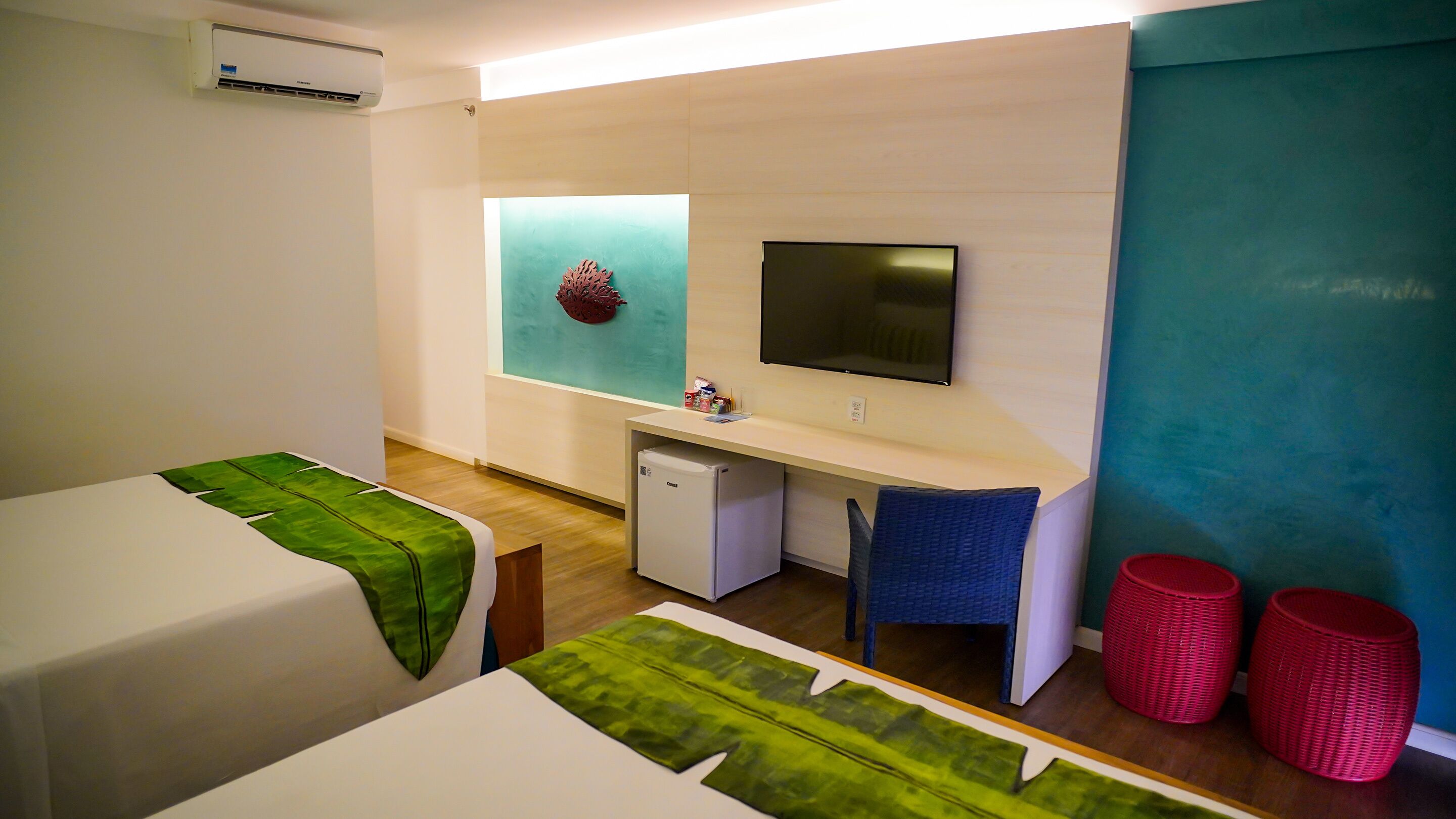 Habitación de hotel con dos camas, un escritorio, un refrigerador pequeño, un televisor y una ventana con un adorno.