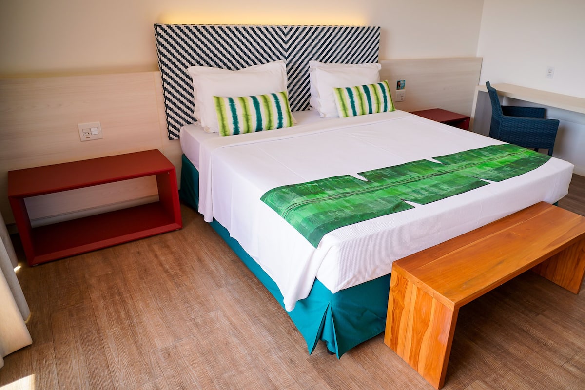 Une chambre bien rangée, avec un lit, un banc, une table de chevet et un petit réfrigérateur.