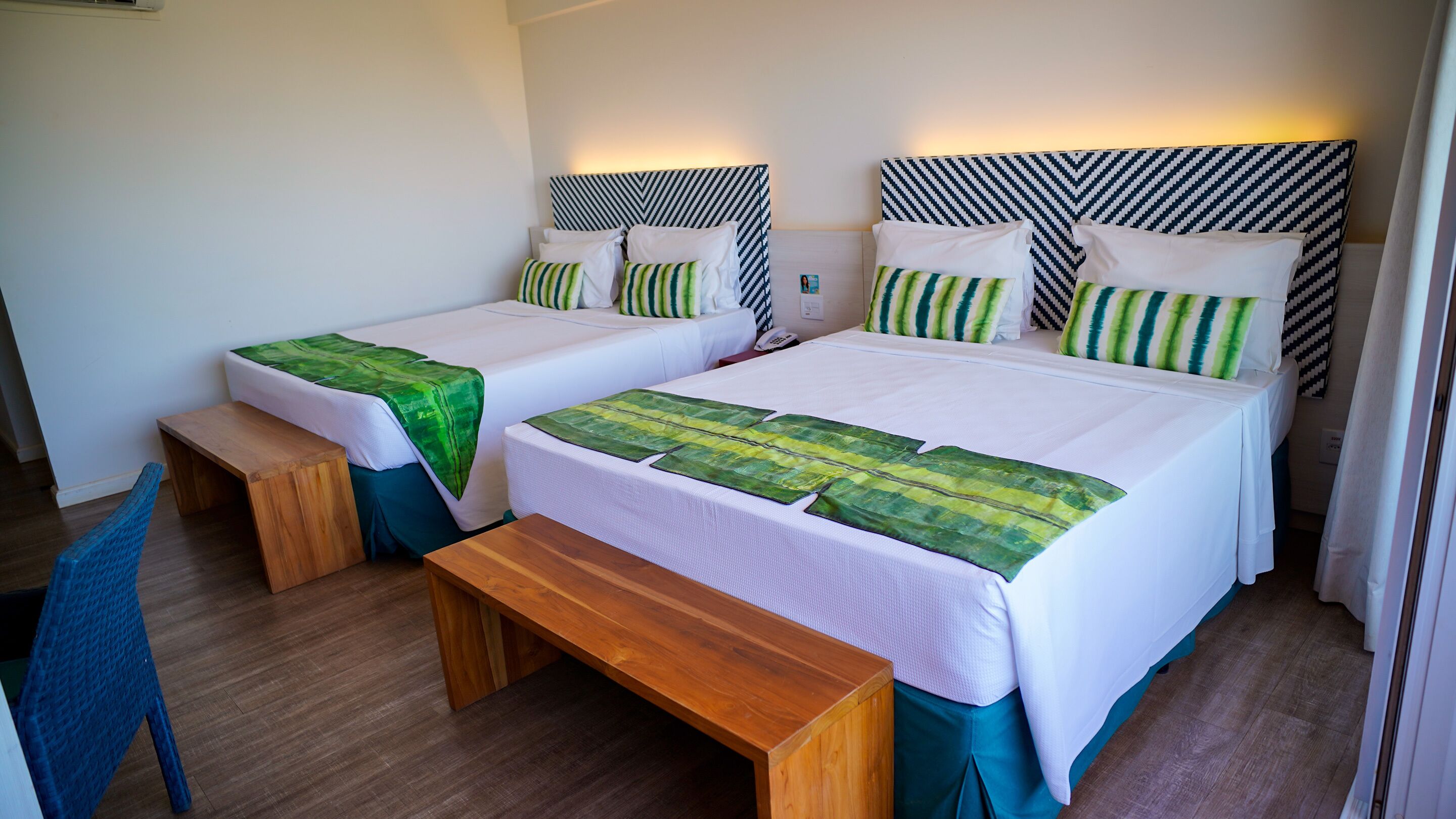 A hotel room with two beds, each with a green leaf-patterned bedspread, a wooden bench, and a patterned headboard.