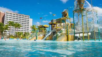 Un parc aquatique avec des toboggans et des jeux d’eau, une piscine, des palmiers et un hôtel en arrière-plan.