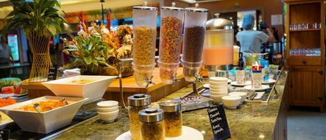 A buffet area with various food dispensers, bowls of cereal, and a sign indicating available items like cereal, chocolate, and granola.