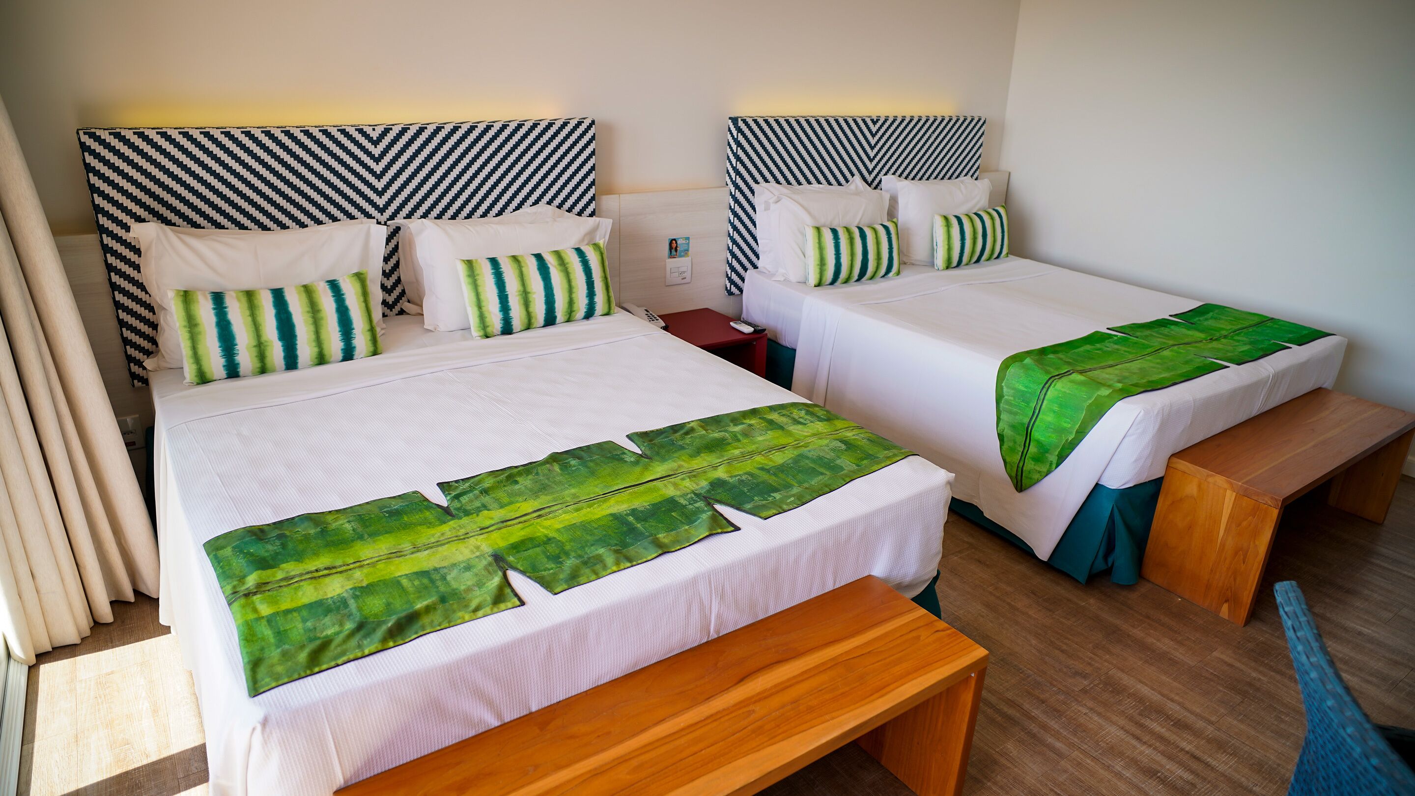 Quarto de hotel com duas camas, cada uma com cabeceira estampada, roupa de cama branca e mantas decoradas com padrão de folhas verdes. Há um banco de madeira e uma pequena mesa com telefone e controle remoto.