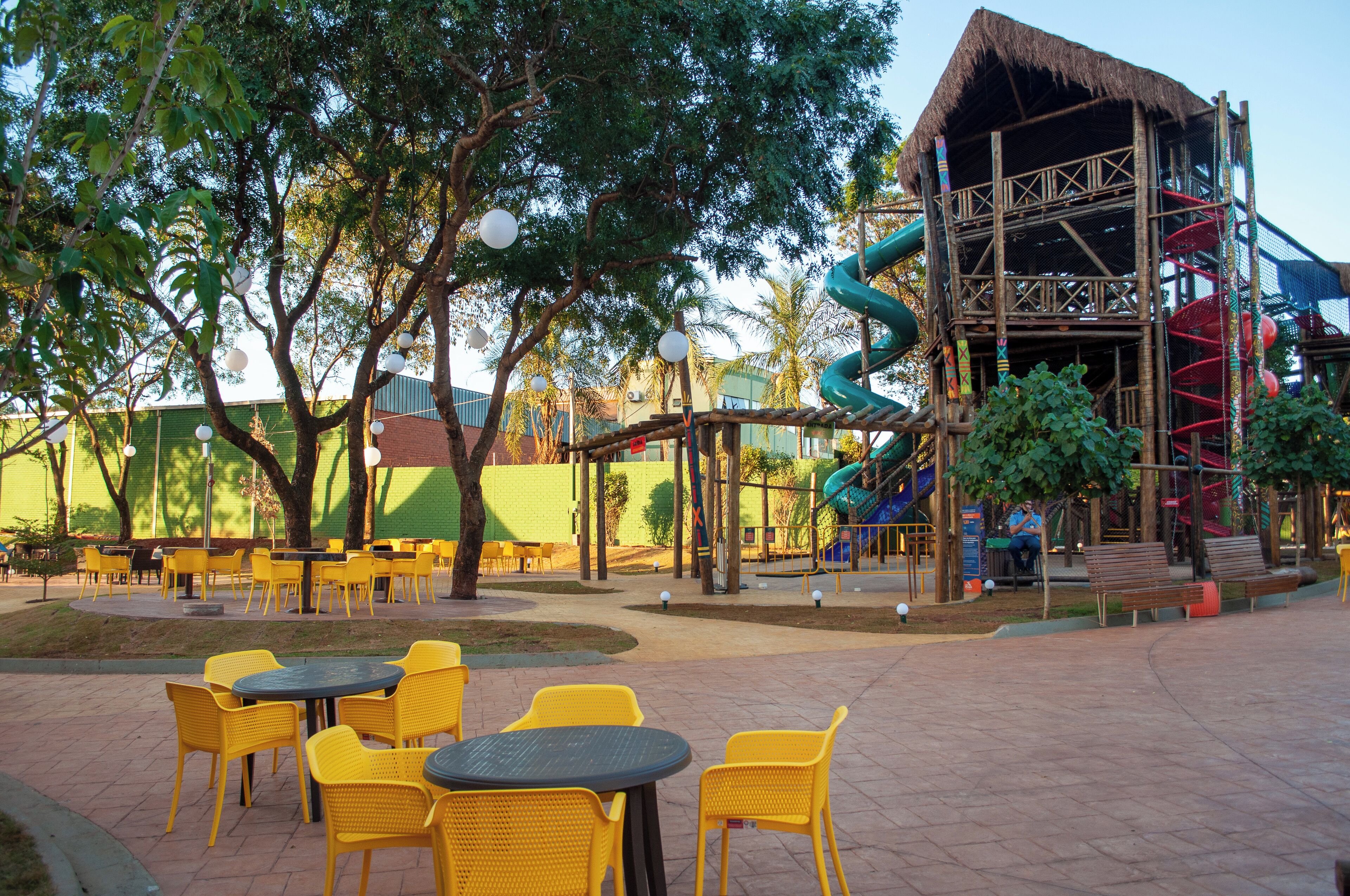 Un espace extérieur avec des chaises et des tables jaunes, une structure de jeux et un bâtiment doté d’un toit de chaume.