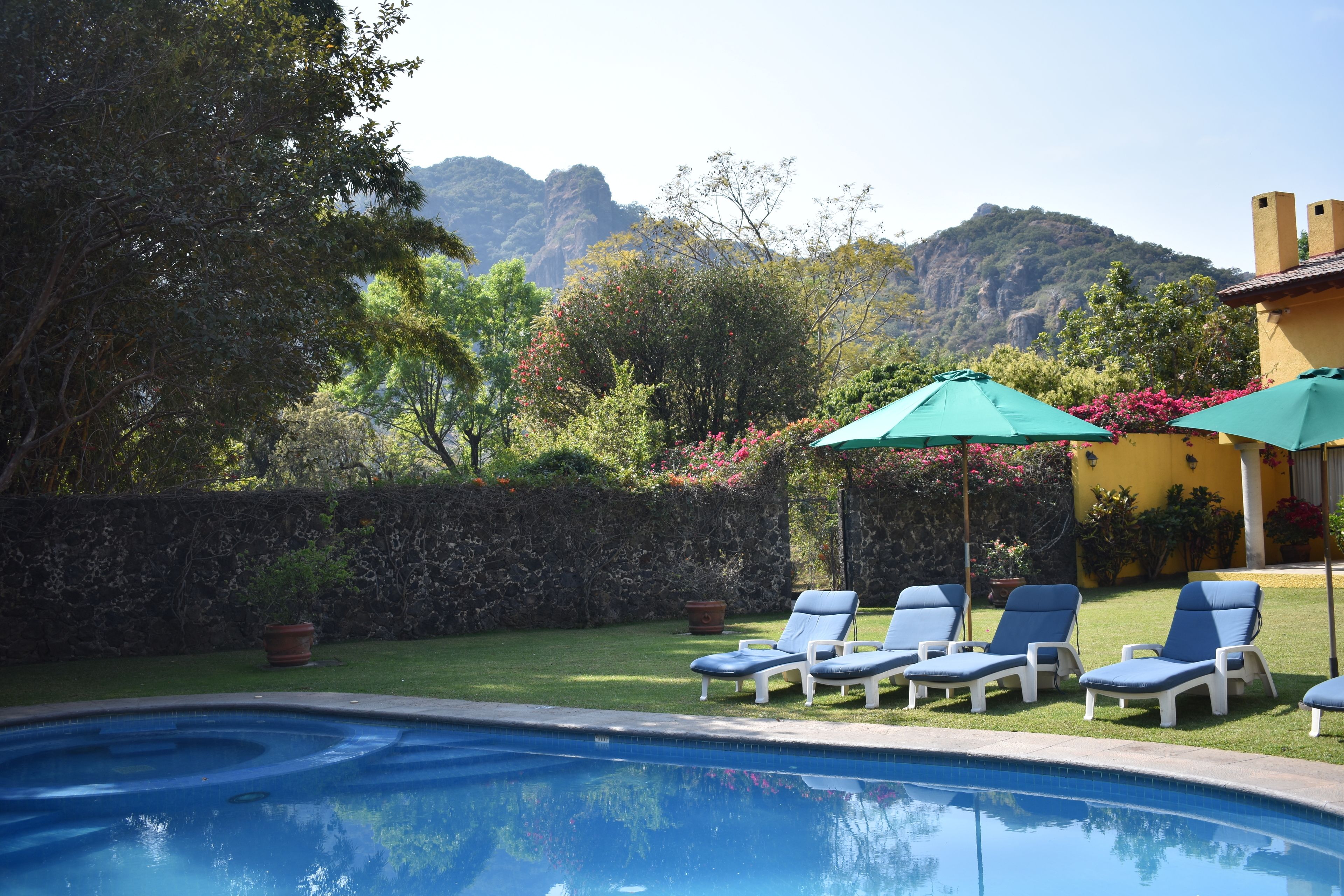 Piscina con sillas de descanso y sombrillas, rodeada de vegetación y con montañas al fondo.