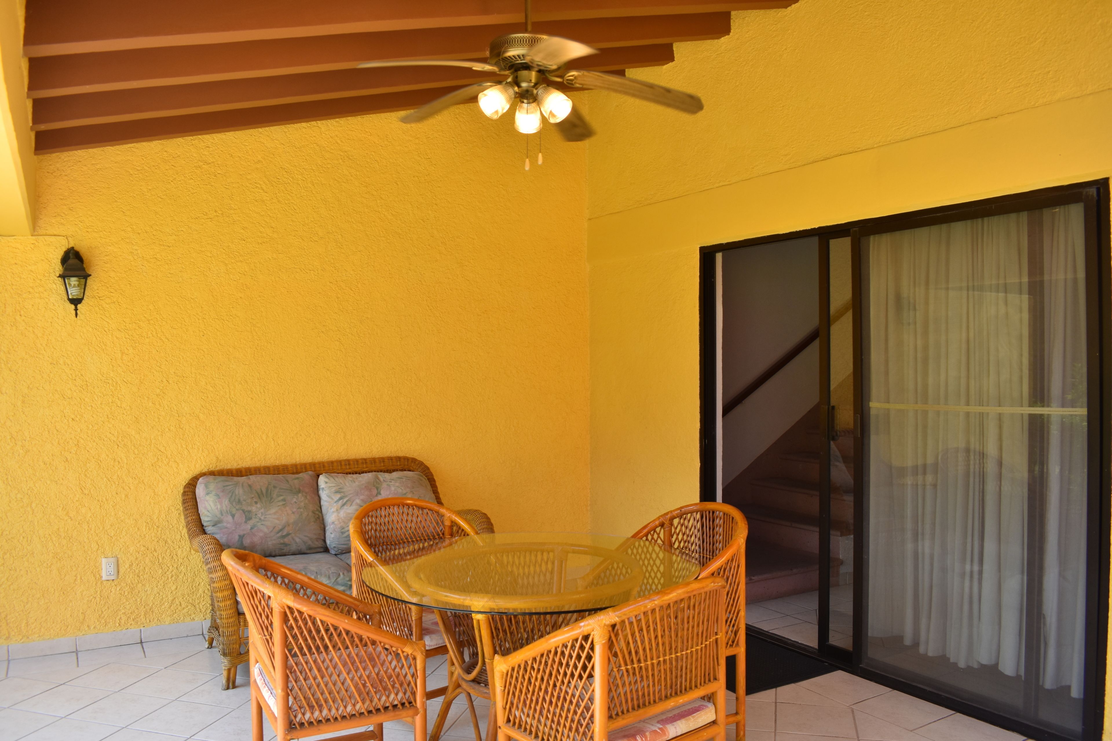 Una habitación con una mesa de vidrio, sillas de mimbre, un ventilador de techo, un sofá y una puerta corredera de cristal.