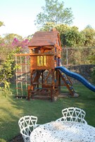 A wooden play structure with a slide and climbing elements, surrounded by a grassy area and a white metal table.