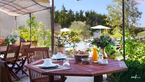 A wooden table set for breakfast with a basket of pastries, a pitcher of orange juice, and a vase with flowers. - Chambres d'hôtes - Les Vents Bleus (Donnazac)