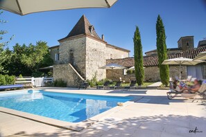 Un edificio de piedra con piscina, sillas de descanso y sombrillas.
