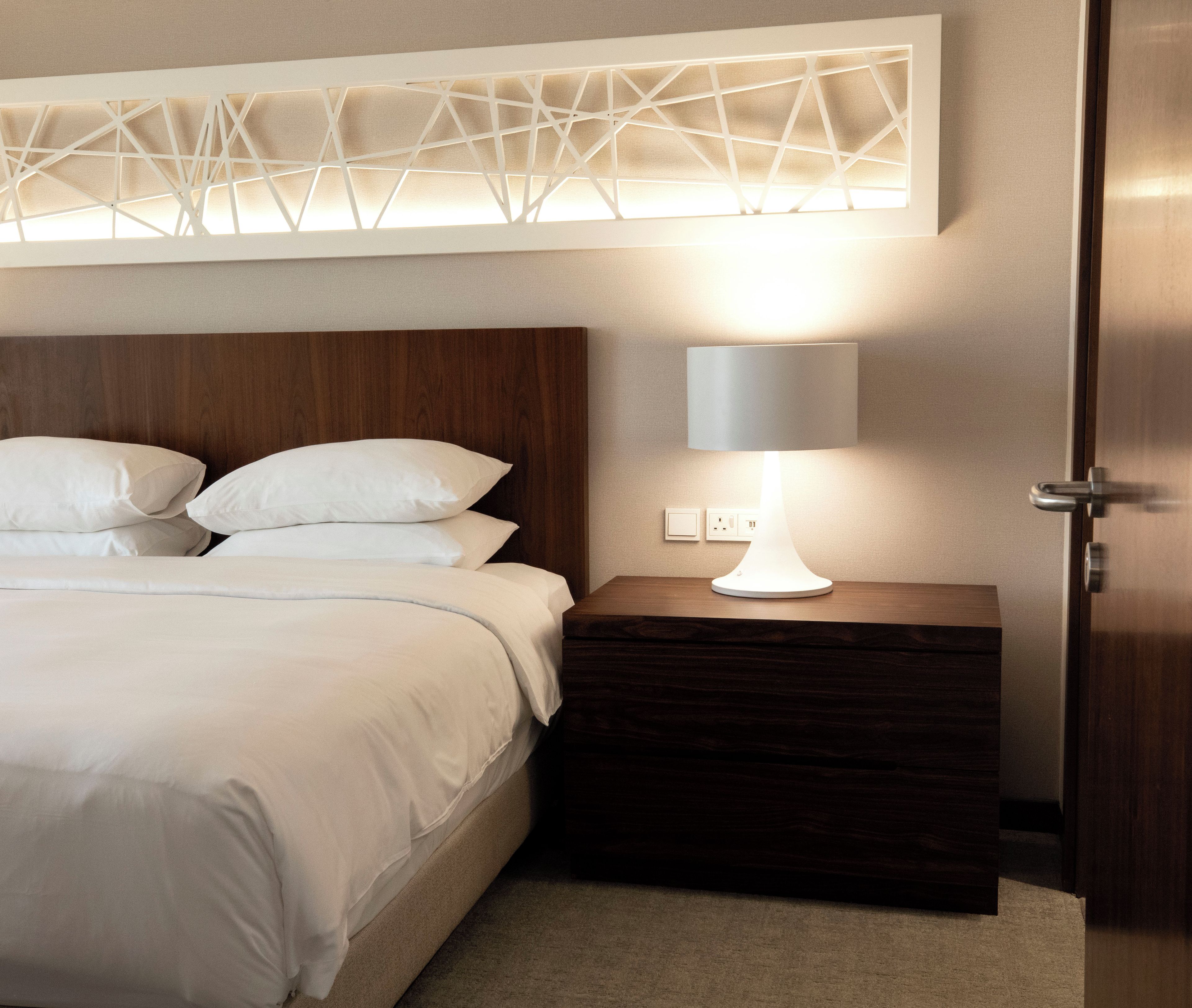 A neatly made bed with white linens, a wooden headboard, a bedside table with a lamp, and a wall-mounted light fixture.