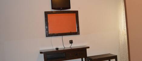 A room with a television mounted on the wall, a wooden desk with a bench, and a patterned cushion.