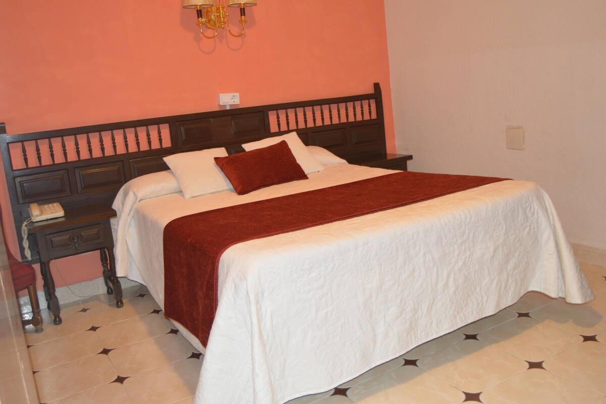 A neatly made bed with a red throw, a bedside table with a telephone, and a wall-mounted lamp.