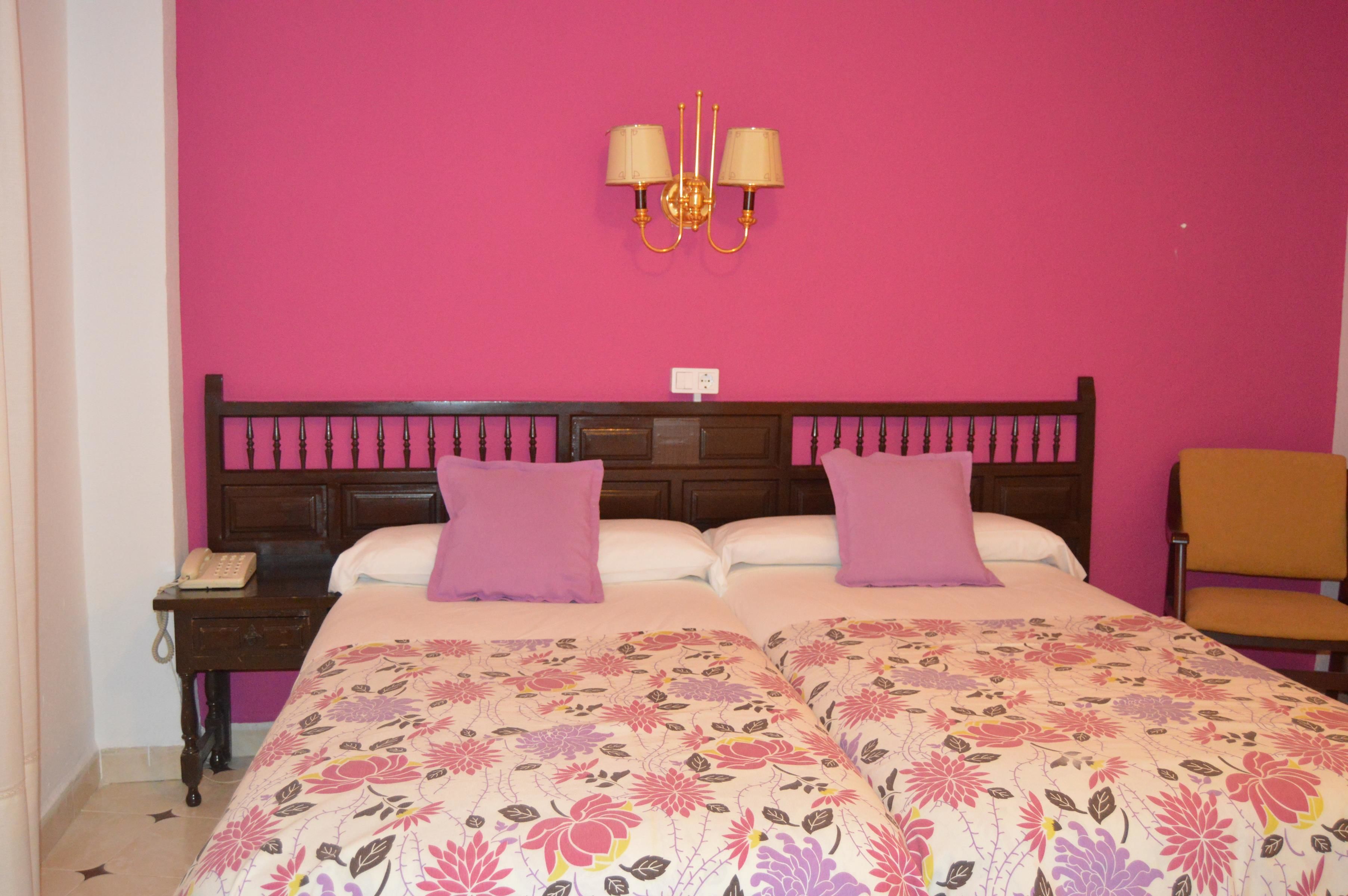 A bedroom with a pink wall, a wooden headboard, two beds with floral bedding, two purple pillows, a nightstand with a telephone, and a chair.