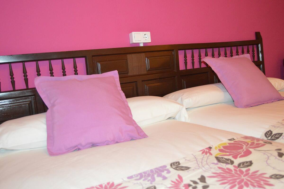 A double bed with a floral bedspread and two pillows, one pink and one white, in a room with a wooden headboard and pink walls.