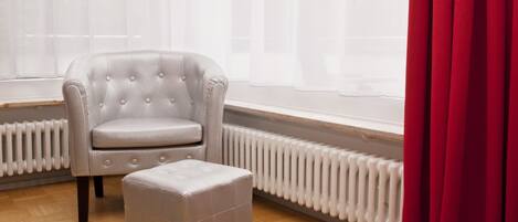 A room with a tufted armchair and ottoman, a radiator, and a window with sheer curtains.