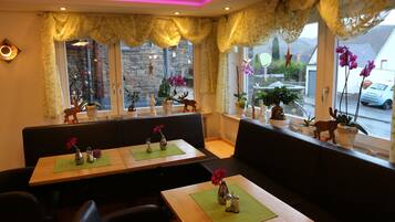 Un restaurante con mesas de madera y asientos tipo banca acolchados en negro, decorado con plantas y flores en macetas.