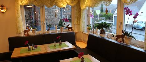 Un restaurante con mesas de madera y asientos tipo banca acolchados en negro, decorado con plantas y flores en macetas.