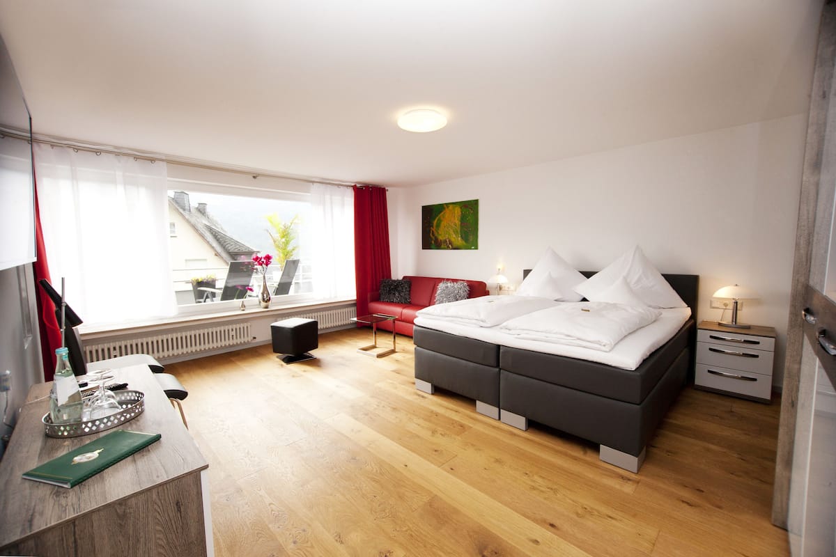 A modern bedroom with a large bed, a desk with a chair, a red sofa, and a window with a view of a building outside.