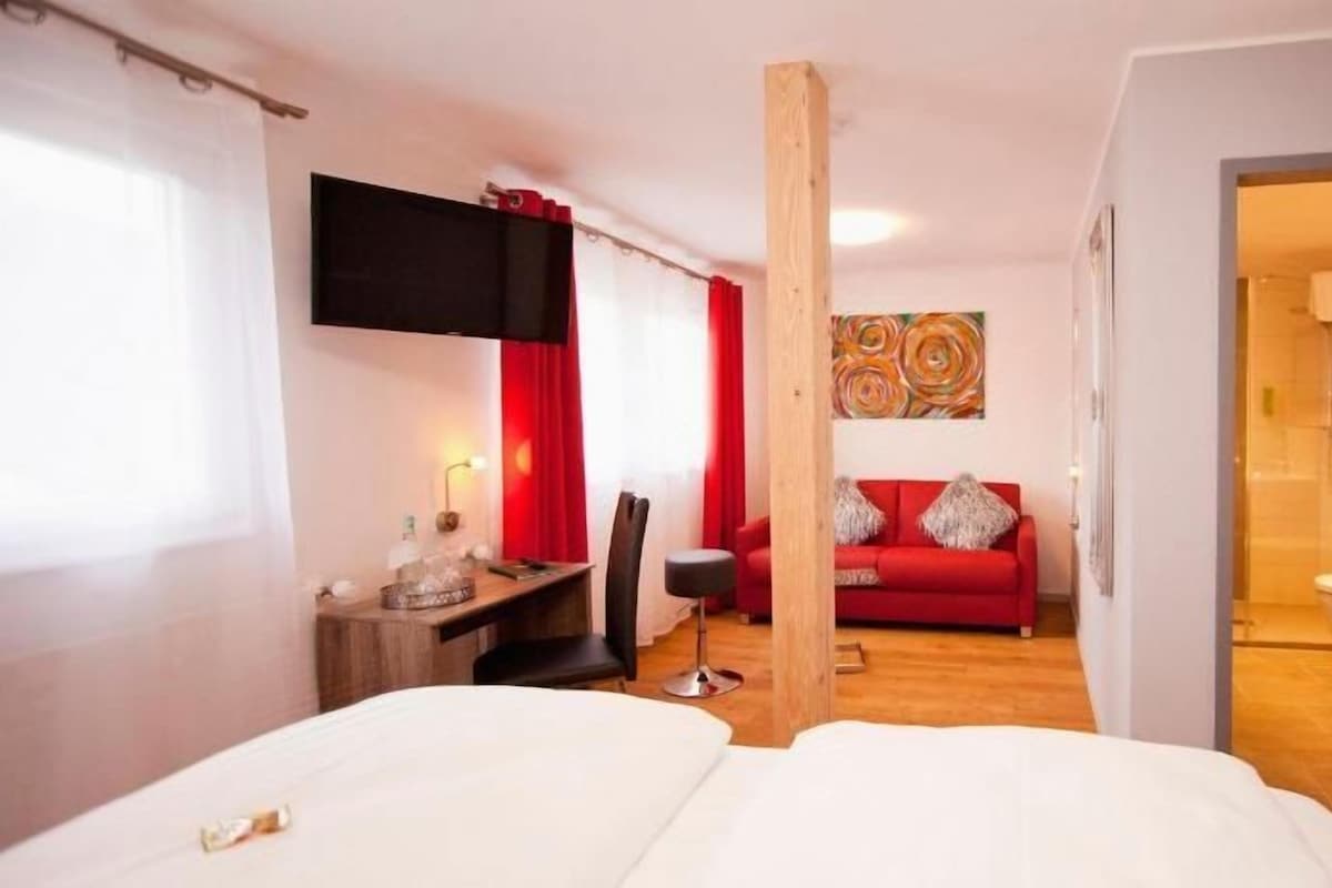 A hotel room with a bed, a desk, a red sofa, and a bathroom visible through an open door.