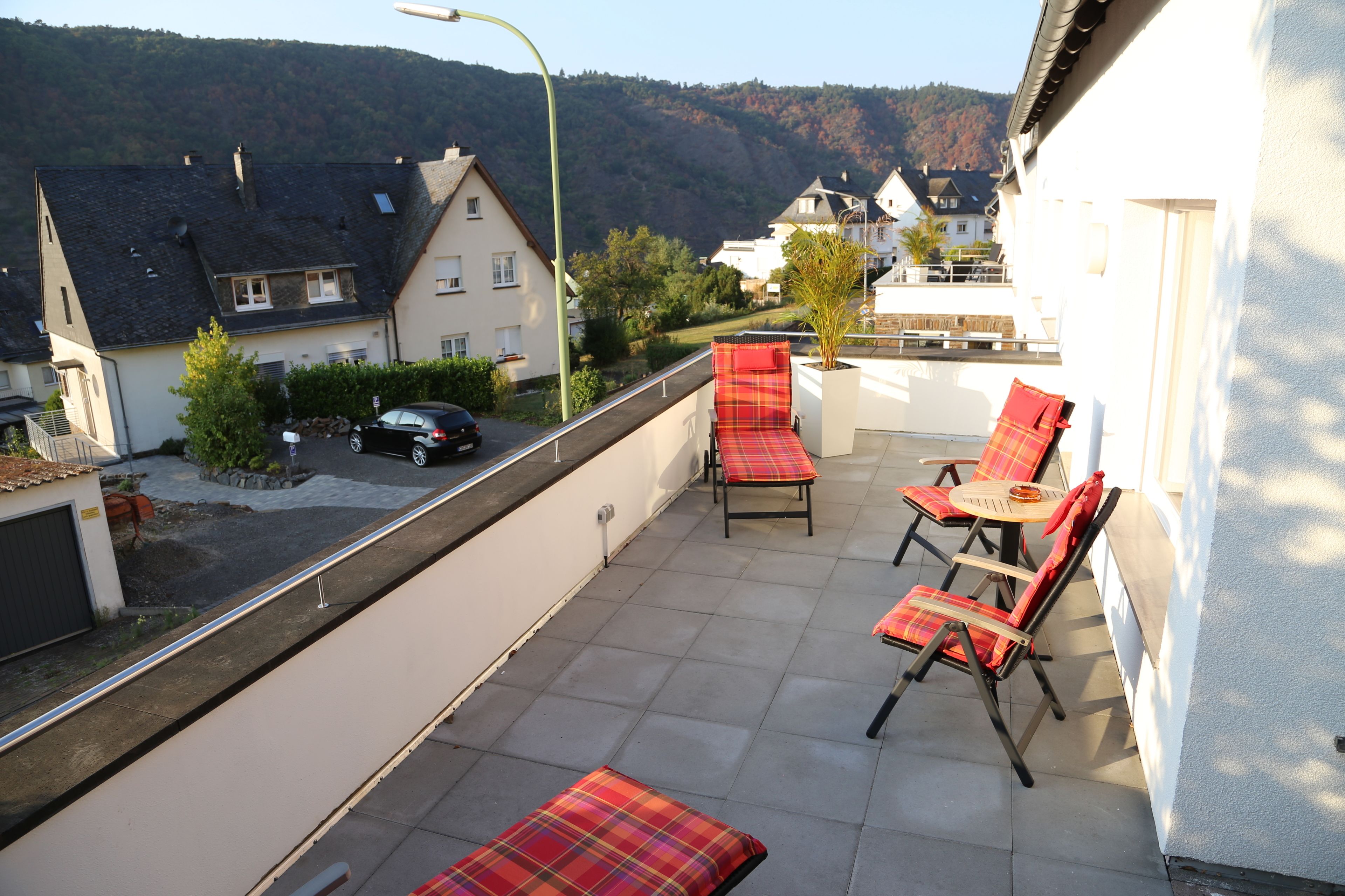A balcony with red plaid chairs and a table, overlooking a street and houses with a mountain in the background.