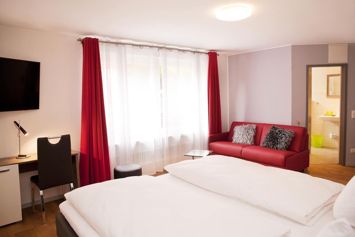 A hotel room with a bed, a desk, a chair, a red sofa, and a bathroom visible through an open door.