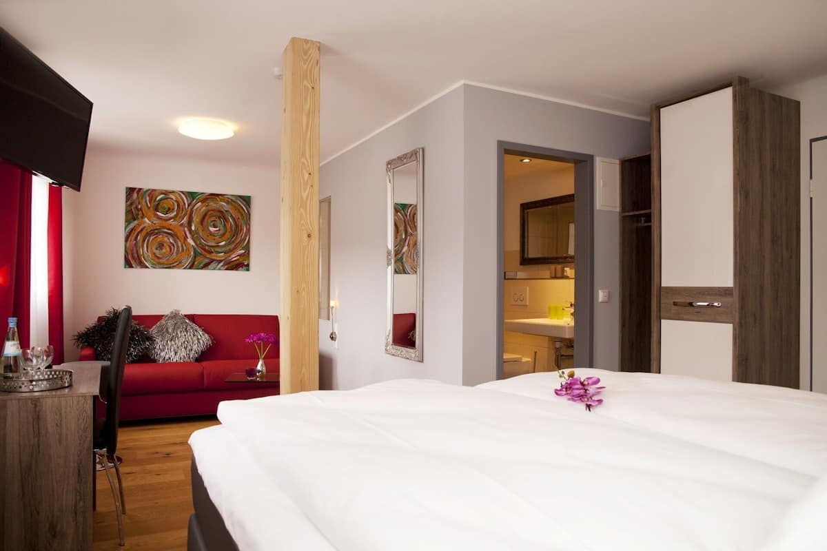 A hotel room with a bed, a red sofa, a wooden pillar, a mirror, and a bathroom visible through an open door.