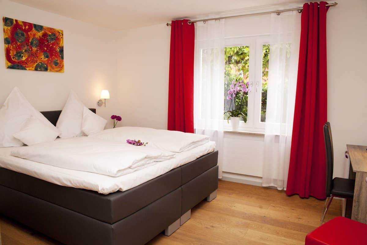 A bedroom with a large bed, red curtains, a window with a view of greenery, and a painting on the wall.