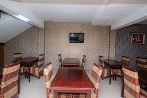 A dining area with a long wooden table and wicker chairs, a television mounted on the wall, and patterned wallpaper. - RedDoorz Plus @ Grand Populer Hotel (Makassar)