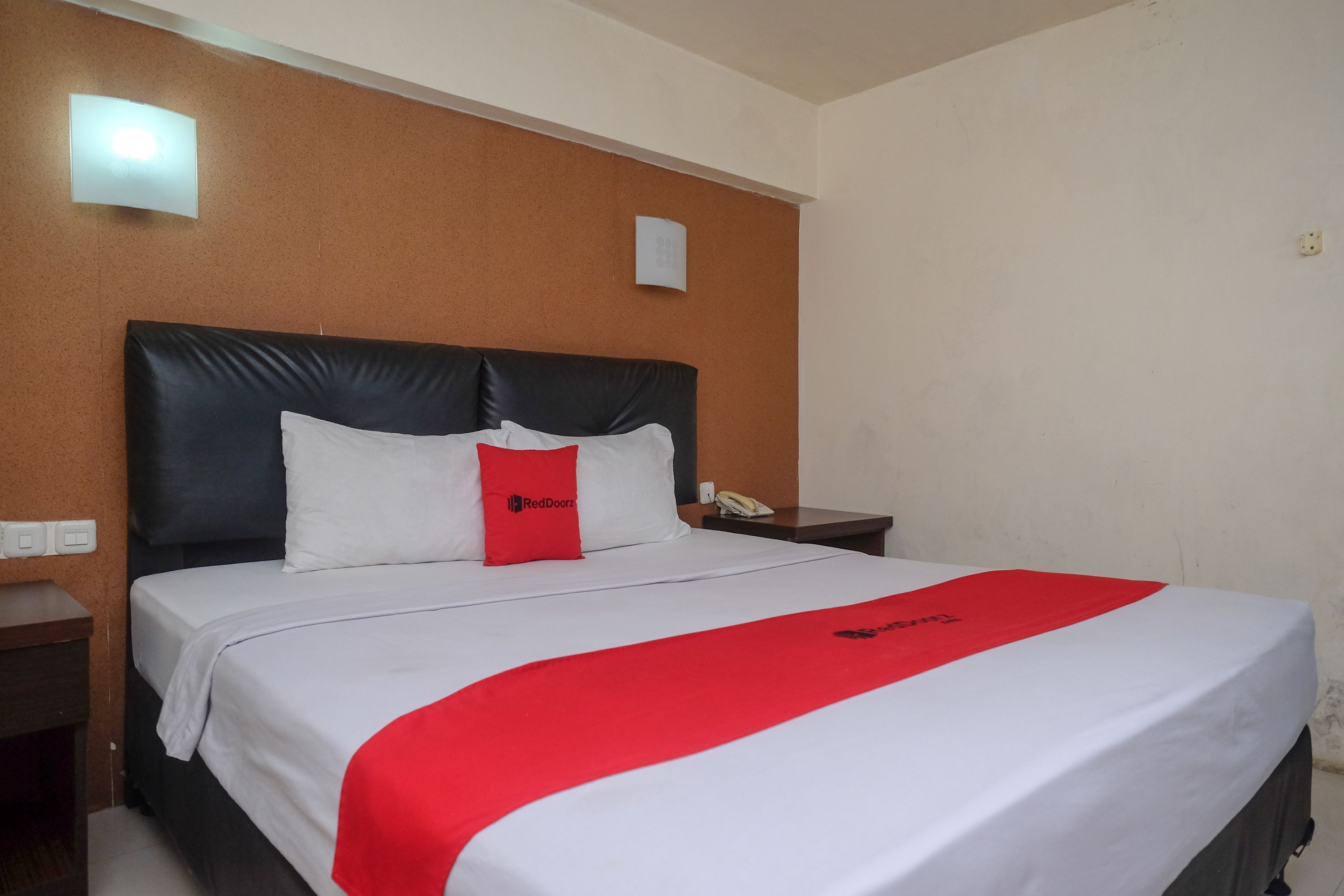 A hotel room with a large bed, a red and white bedspread, a black headboard, and a nightstand with a phone.