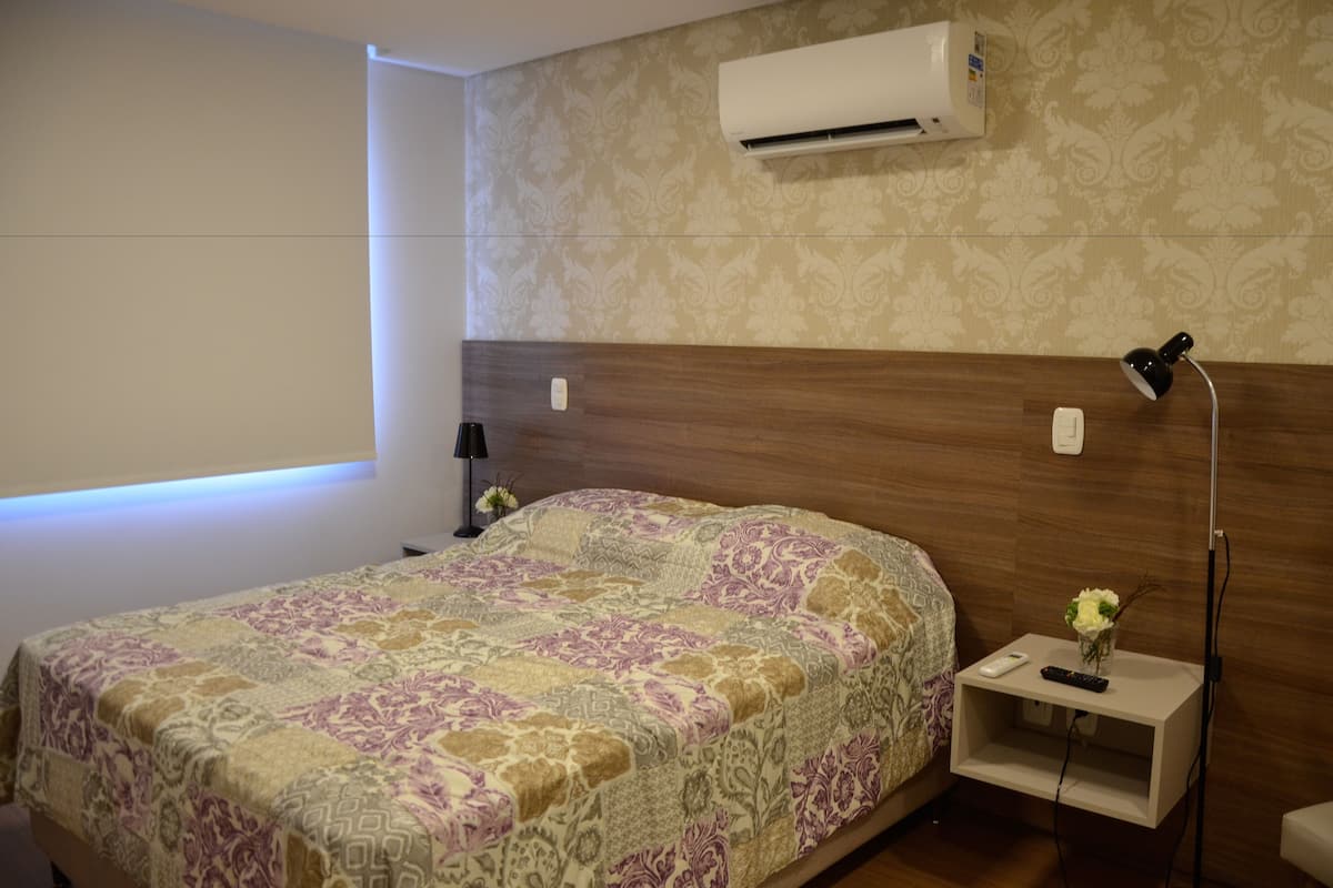 A hotel room with a large bed, an air conditioner, a bedside lamp, a nightstand with a remote control, and a small vase with flowers.