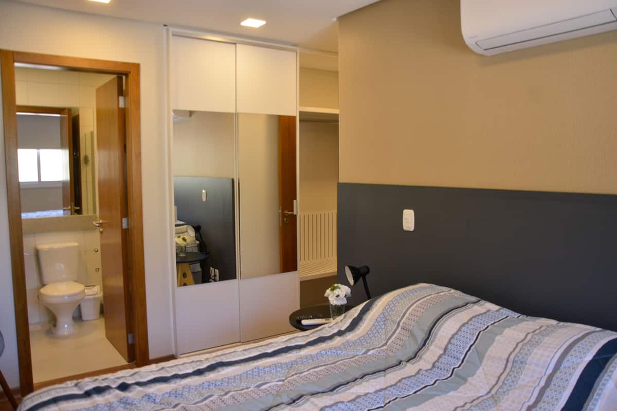 A bedroom with a bed, a bedside table, a mirror, a wardrobe, and a bathroom visible through an open door.