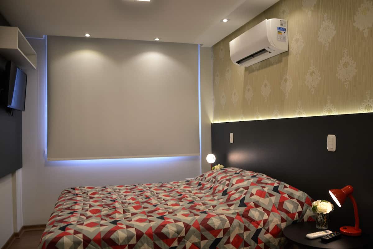 A bedroom with a bed, a bedside table with a lamp, a wall-mounted air conditioner, and a TV on the wall.
