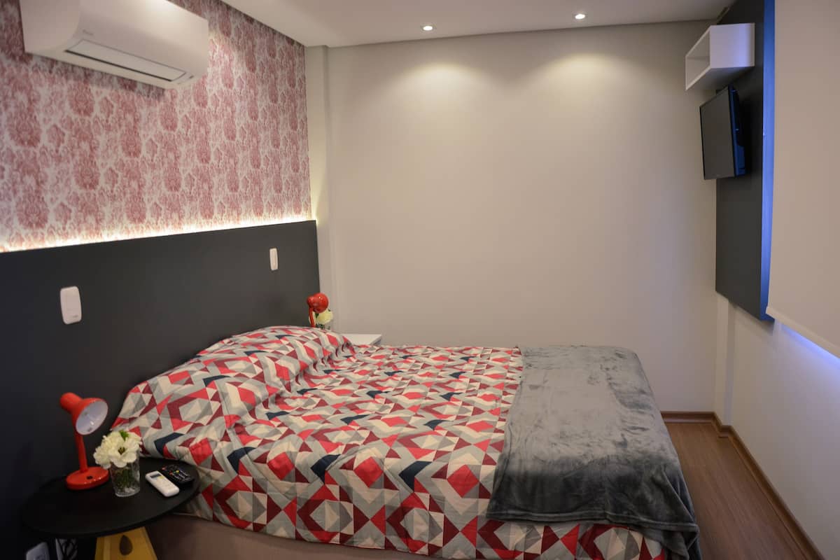 A bedroom with a patterned bedspread, a bedside table with a lamp and remote controls, and a wall-mounted TV.